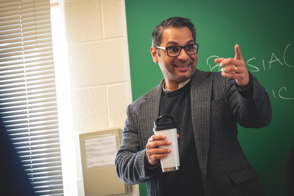 A man in glasses and a blazer holding a travel mug gestures and smiles while standing in front of a green chalkboard in a classroom.