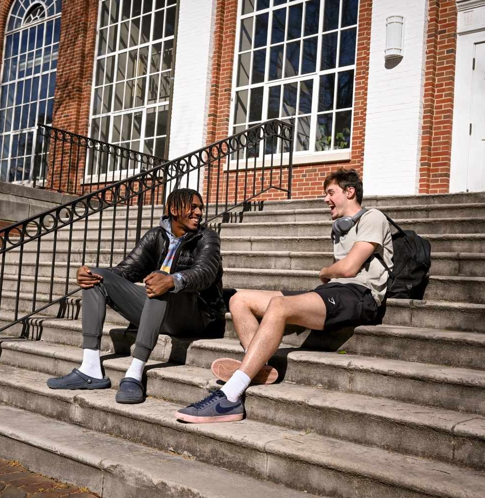 52768896349_832cf76f73_o Two young men sit on outdoor steps in front of a brick building, talking and laughing. One wears a jacket and pants; the other wears a T-shirt, shorts, and has headphones around his neck.