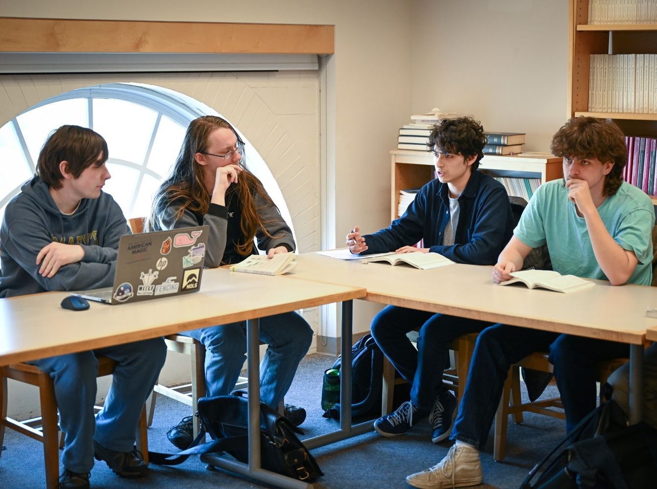 52769050940_a688e56c25_o Four students sit around a table in a library or classroom, discussing books open in front of them; one has a laptop with stickers on it.