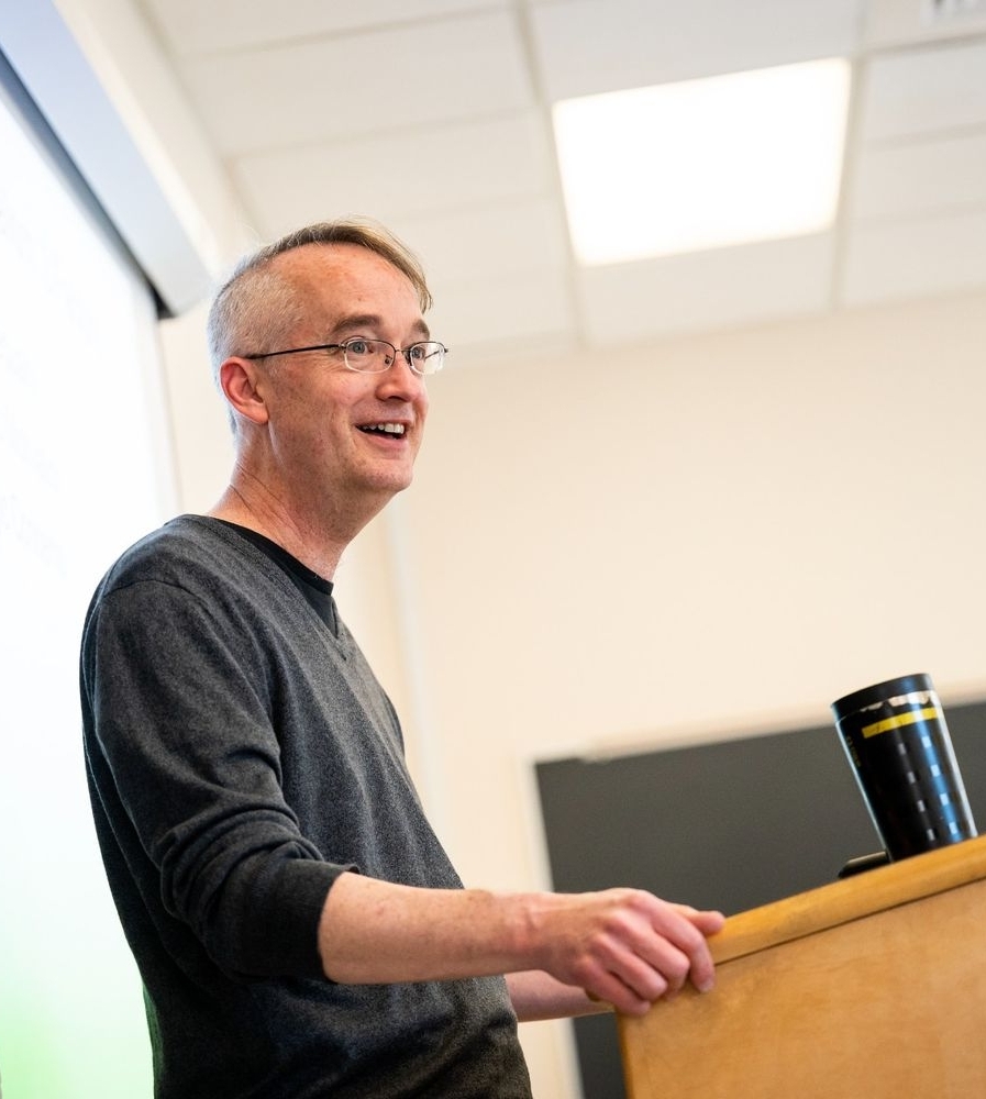 52769125313_464b3625a4_o A man wearing glasses stands at a lectern, speaking in a classroom with a projection screen and chalkboard in the background.