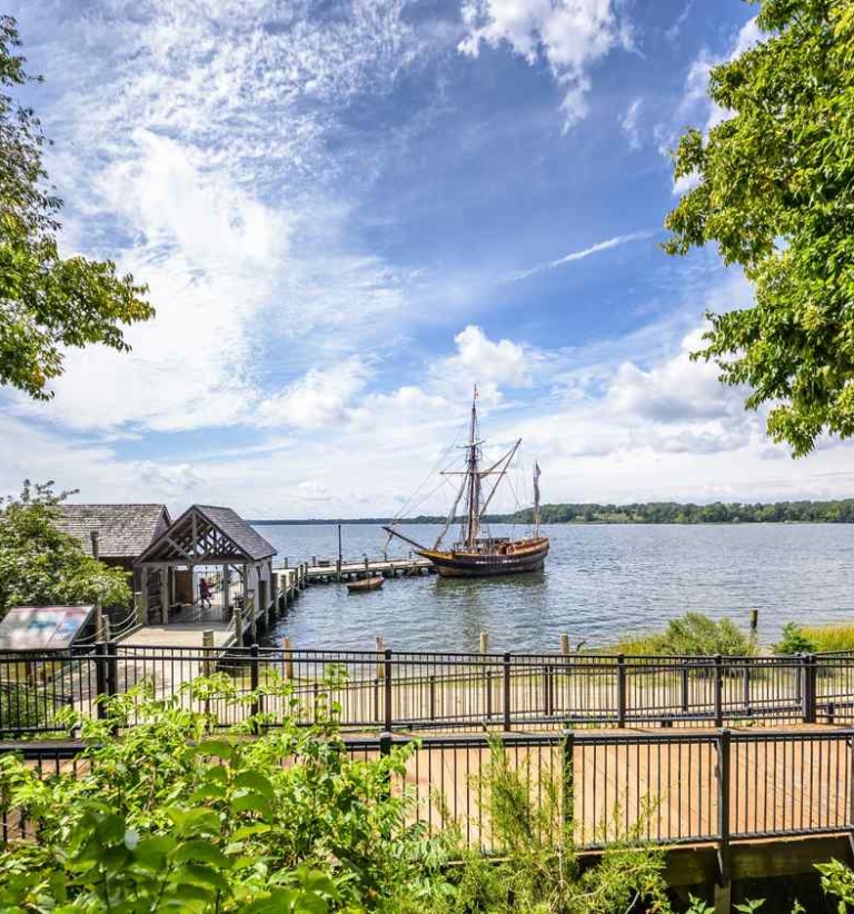 53979969968_44b4417339_o A wooden sailing ship is docked at a pier on a wide river, with trees, a footbridge, and a small building in the foreground under a partly cloudy sky.