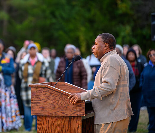 A man speaks at a wooden podium outdoors to a diverse crowd standing on grass, with trees visible in the background.
