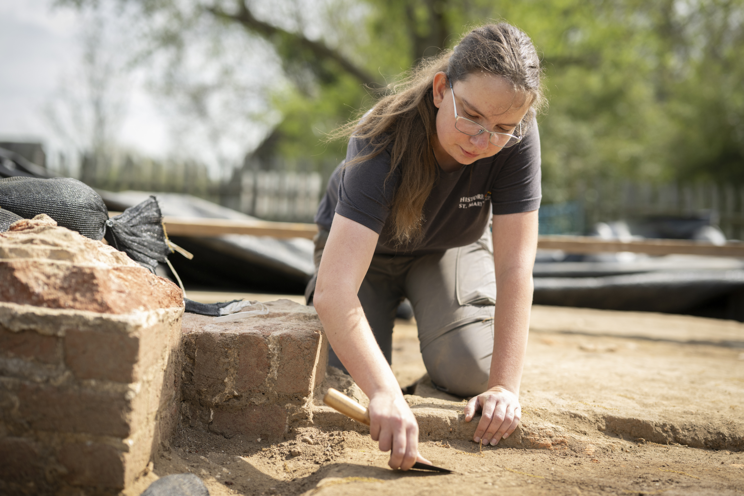54540505795_7e639618a0_o A person kneels on the ground, carefully using a small tool to excavate soil at an outdoor archaeological site.