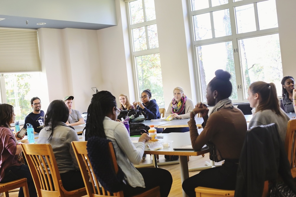 14@SaintMary’sCollege_0027 A group of people sit around a large table in a bright room with large windows, engaged in discussion.