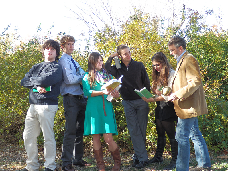Six people stand outdoors in front of green foliage, holding books and interacting with each other, appearing engaged in conversation or study.