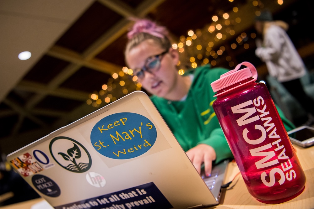A person works on a laptop with various stickers on it; a pink SMCM Seahawks water bottle sits in the foreground.
