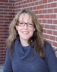 A woman with long, light brown hair and glasses is smiling while sitting in front of a brick wall, wearing a gray cowl-neck sweater.