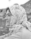 A woman in profile wearing a patterned headscarf sits outdoors, with a rocky background visible behind her.