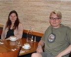 Two people sit at a table in a cafe with cups, glasses, and a water bottle in front of them.