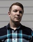 A person with short brown hair wearing a plaid shirt stands in front of a light-colored horizontal siding wall.