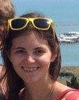 A woman with long brown hair and yellow sunglasses on her head smiles at the camera with a body of water and a pier in the background.