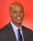 A man in a gray suit, blue striped tie, and light blue shirt smiles at the camera against a red background.