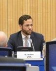 A man in a suit sits at a conference table with a nameplate reading "R. Akbar, USA" in front of him, speaking at a formal event.