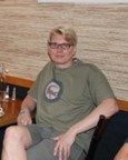 A person with short blonde hair and glasses sits at a table, wearing a green t-shirt and shorts, with one hand resting on the table and the other on their lap.