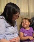 An adult and a young girl sitting on a couch, facing each other. The girl is smiling at the adult, who is looking down at her.