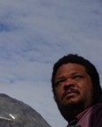 A man with short hair and glasses looks into the distance outdoors under a cloudy sky.