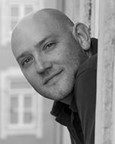 A man with a shaved head leans out of a window, looking at the camera with a slight smile. The photo is in black and white.