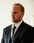 A man with short blond hair and a beard wears a black suit and tie, standing indoors against a light background.