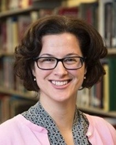 A woman with short brown hair and glasses smiles while standing in front of bookshelves in a library.