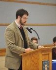 A man in a tan blazer speaks at a podium with a microphone in a conference room; two people are seated in the background.