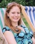 A woman with long red hair sits on a striped deck chair outdoors, smiling at the camera. She is wearing a patterned top and sunglasses on her head.