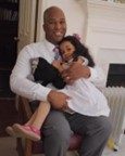 A man in a white shirt and tie sits on a chair, smiling and hugging a young girl in a white dress and pink shoes.