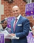 A man in a suit stands at a podium with a microphone, holding papers. People behind him hold signs that say "Robert White." Brick building in the background.