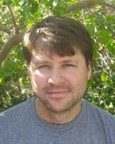 A man with short brown hair and a beard wearing a gray T-shirt stands outdoors in front of green leafy trees.