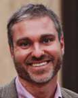 A man with short gray hair and a beard is smiling, wearing a light purple shirt and a brown blazer.