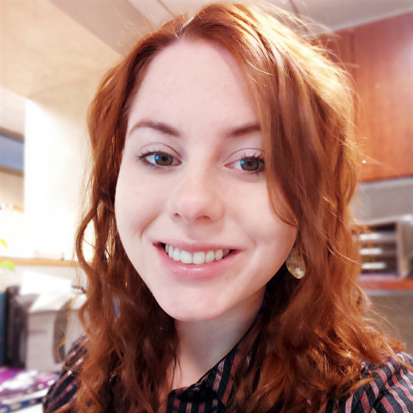 A woman with wavy red hair and fair skin smiles at the camera in a well-lit indoor setting with wooden cabinets in the background.