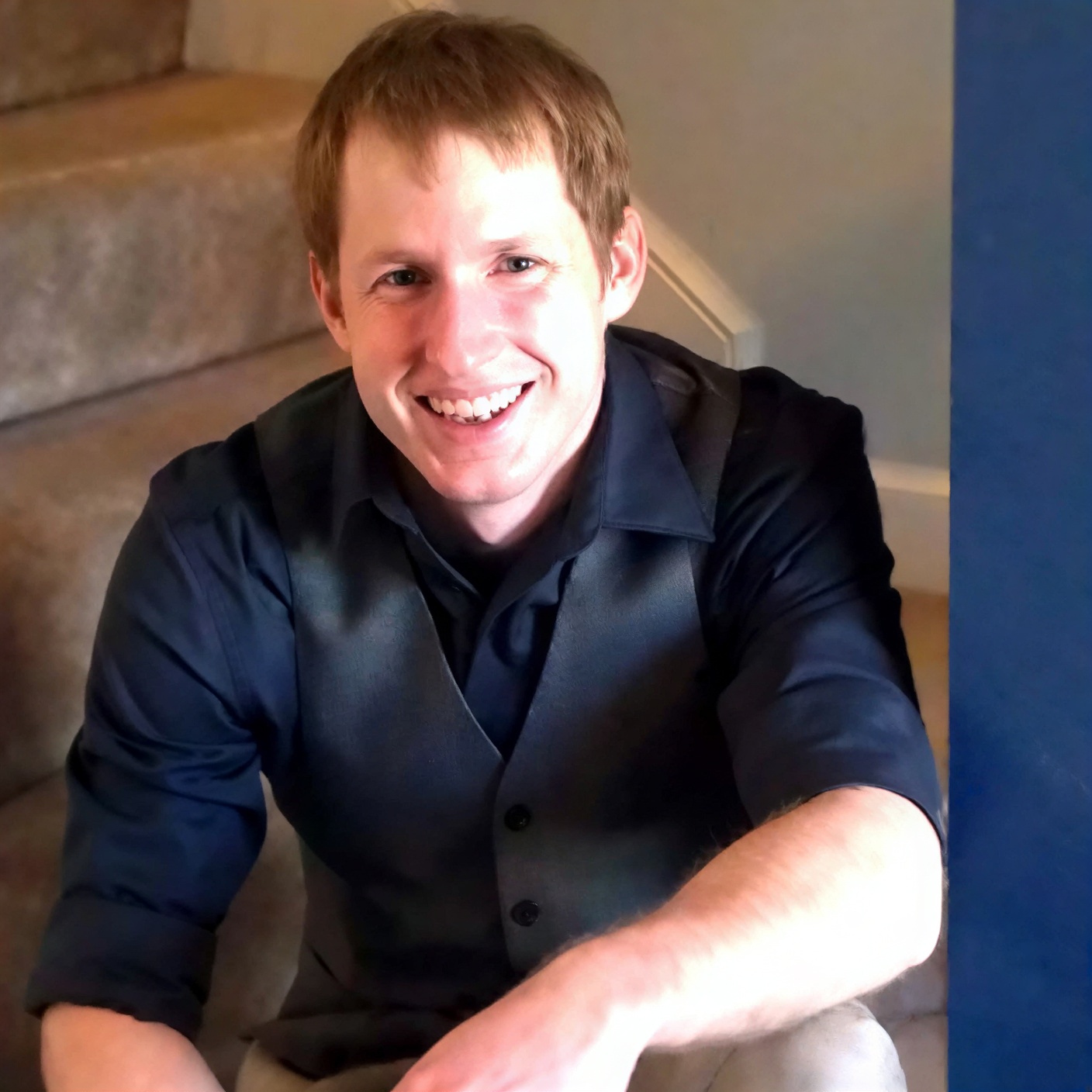 A man with short light brown hair, wearing a black shirt and dark vest, sits on carpeted stairs, smiling at the camera.