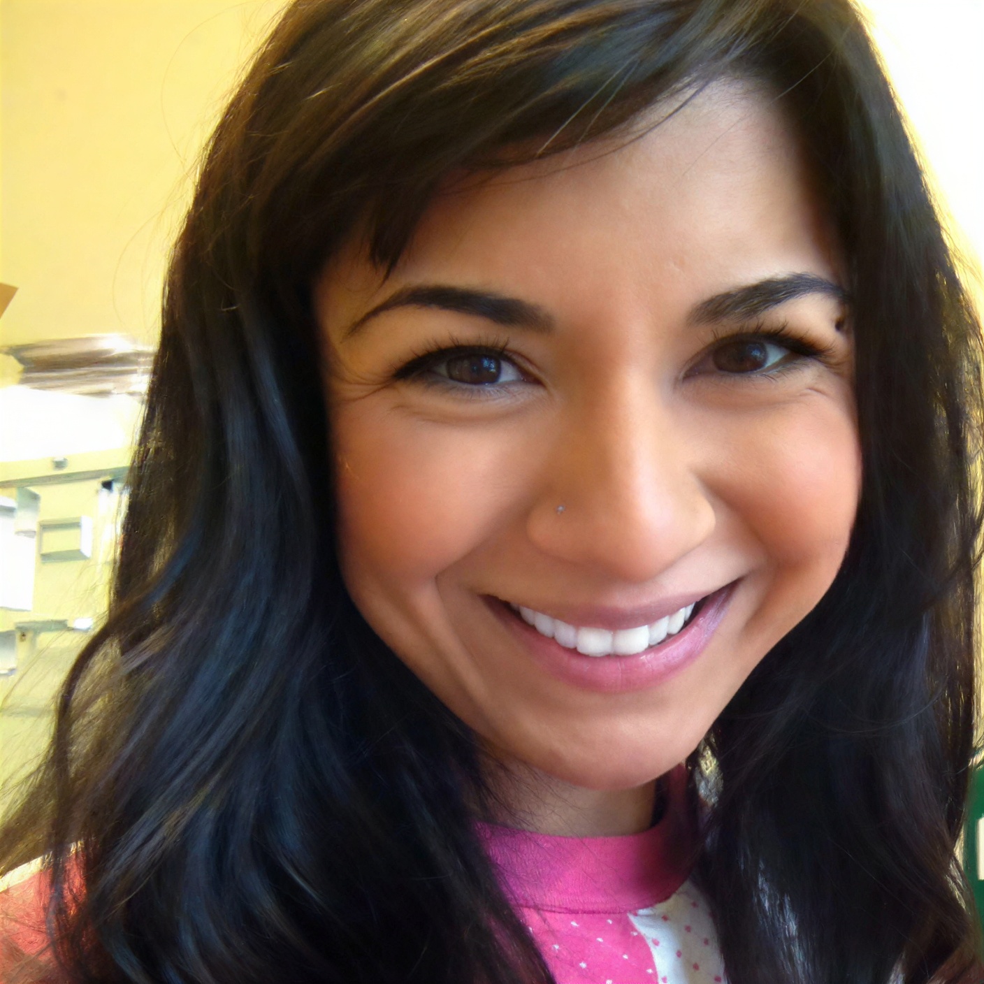 A woman with long dark hair and a nose stud smiles at the camera in an indoor setting.