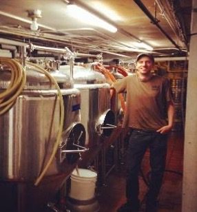 A person stands next to large stainless steel brewing tanks in a brewery or industrial facility.