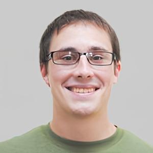 A young adult wearing glasses and a green shirt smiles at the camera against a plain, light gray background.
