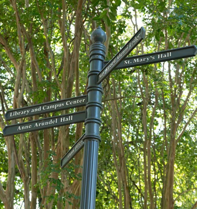 44458783390_f85a01cf04_o A black signpost with directional signs for Library and Campus Center, Anne Arundel Hall, and St. Mary's Hall, surrounded by green trees.