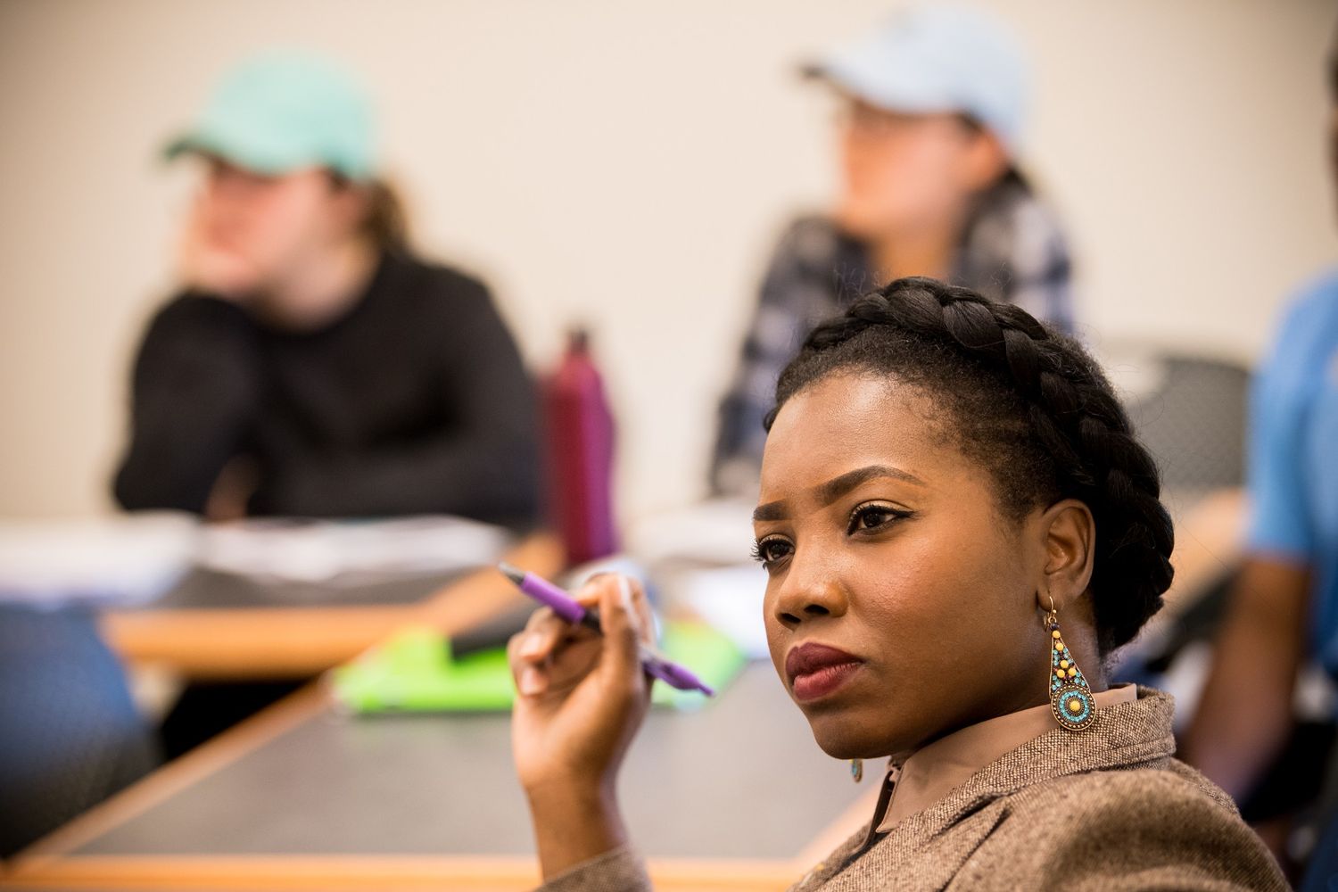 A woman holding a pen and listening attentively in a classroom, with other students seated and slightly out of focus in the background.