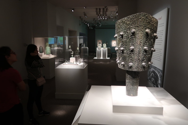 Visitors observe ancient bronze artifacts displayed in glass cases and on pedestals in a dimly lit museum gallery.