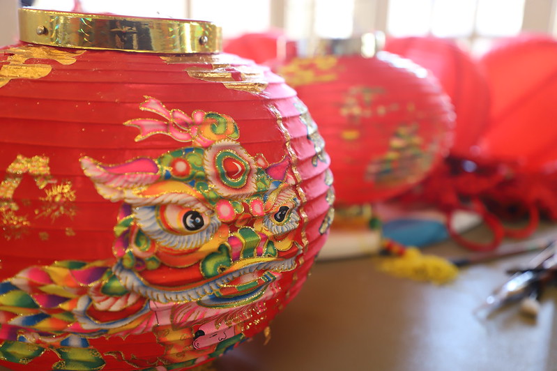 Close-up of a red paper lantern with a colorful dragon design, with more red lanterns and decorations blurred in the background.