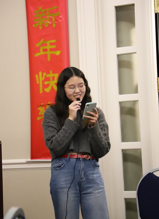 53529778080_162057ffa7_c A young woman speaks into a microphone while reading from her phone, standing next to a red banner with Chinese characters.