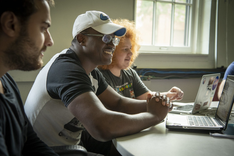 Three people sit at a table with laptops, chatting and smiling in a well-lit room with a window in the background.