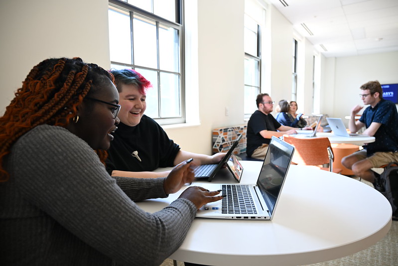 Two people sit at a round table working on laptops, with a group of others using laptops at a table in the background in a bright, modern room.