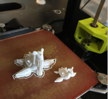 Two partially finished white 3D printed models sit on a printer bed, with one appearing incomplete or detached; parts of the 3D printer are visible in the background.