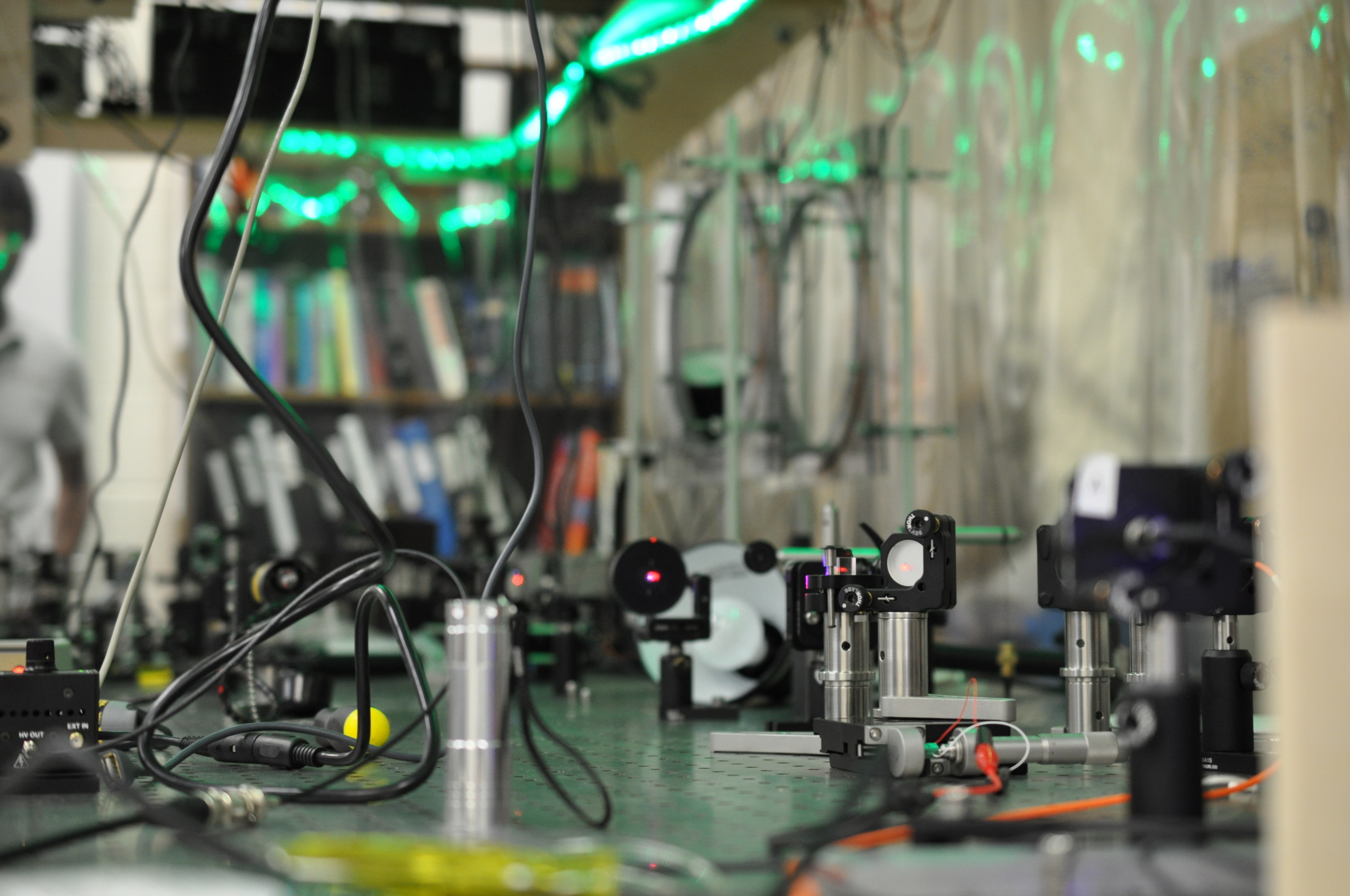 DSC_0682 A laboratory optical table with various scientific instruments, lenses, and wires, with green LED lights and shelves of books in the background.