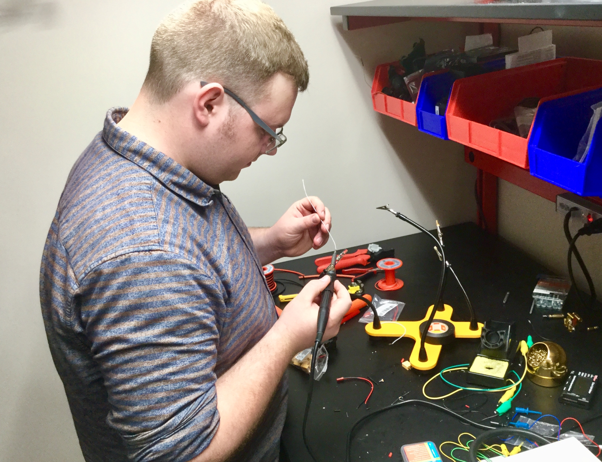 David Gerrish UAV intern2 A person soldering wires at a workbench with electronic components, tools, and parts organized in bins and trays.
