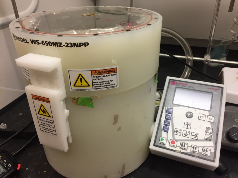 A laboratory spin coater labeled "MODEL WS-650MZ-23NPP" sits next to a control panel; warning stickers indicate chemical splash and pinch point hazards.