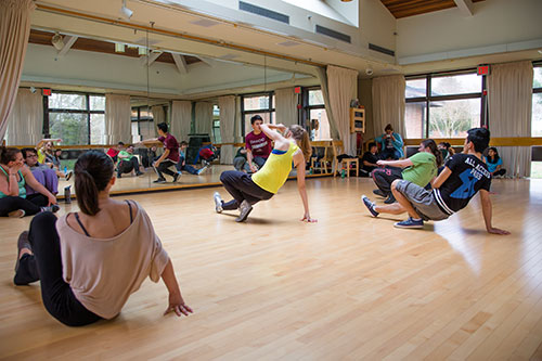 A group of people practice dance moves in a mirrored studio with wooden floors and large windows.