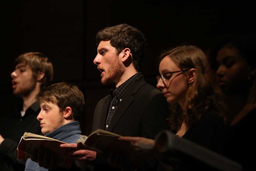 A group of people holding open books sing together in a dimly lit setting.