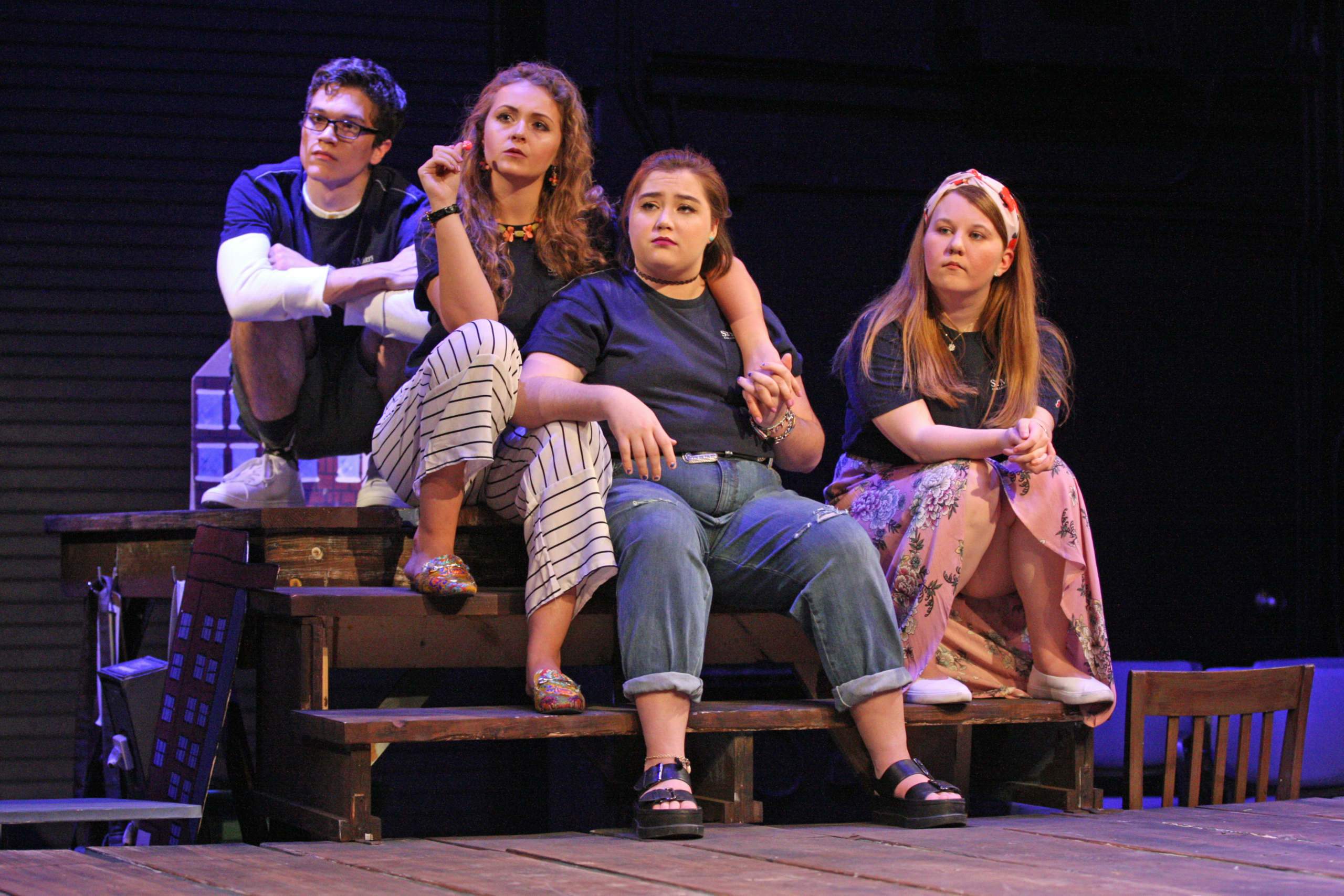 41790462252_d0ee05ab00_o Four people sit closely together on a wooden stage set, wearing casual outfits, and look in various directions with thoughtful or serious expressions.