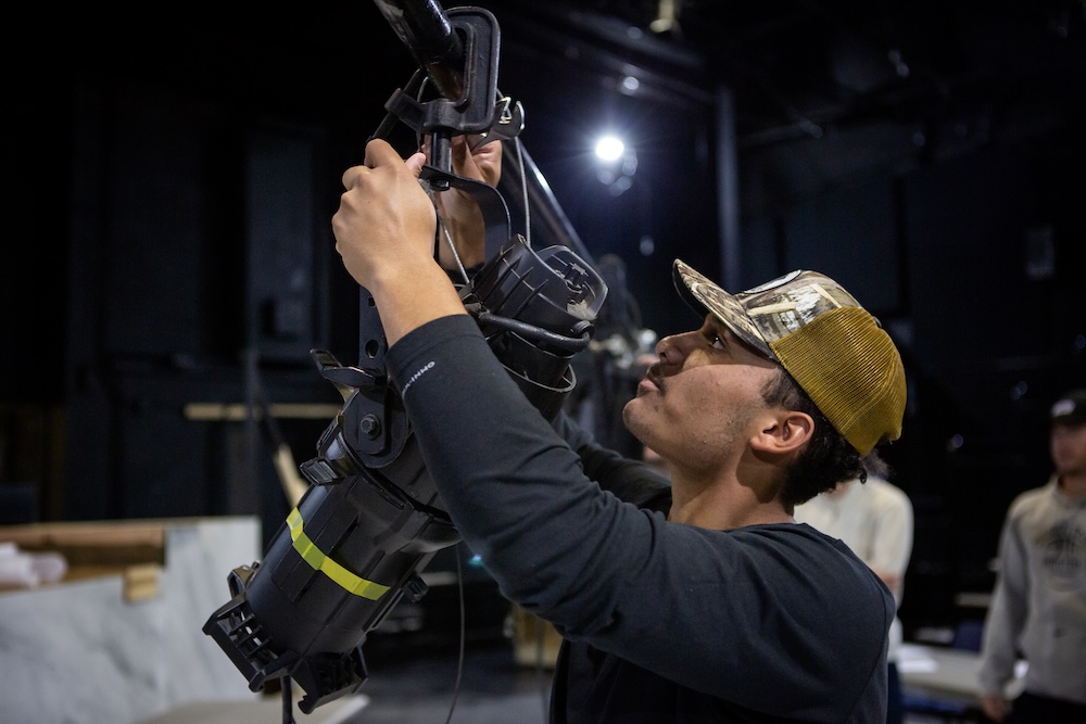 A person in a cap adjusts a stage light fixture in a dark indoor setting, focusing on positioning the equipment overhead.