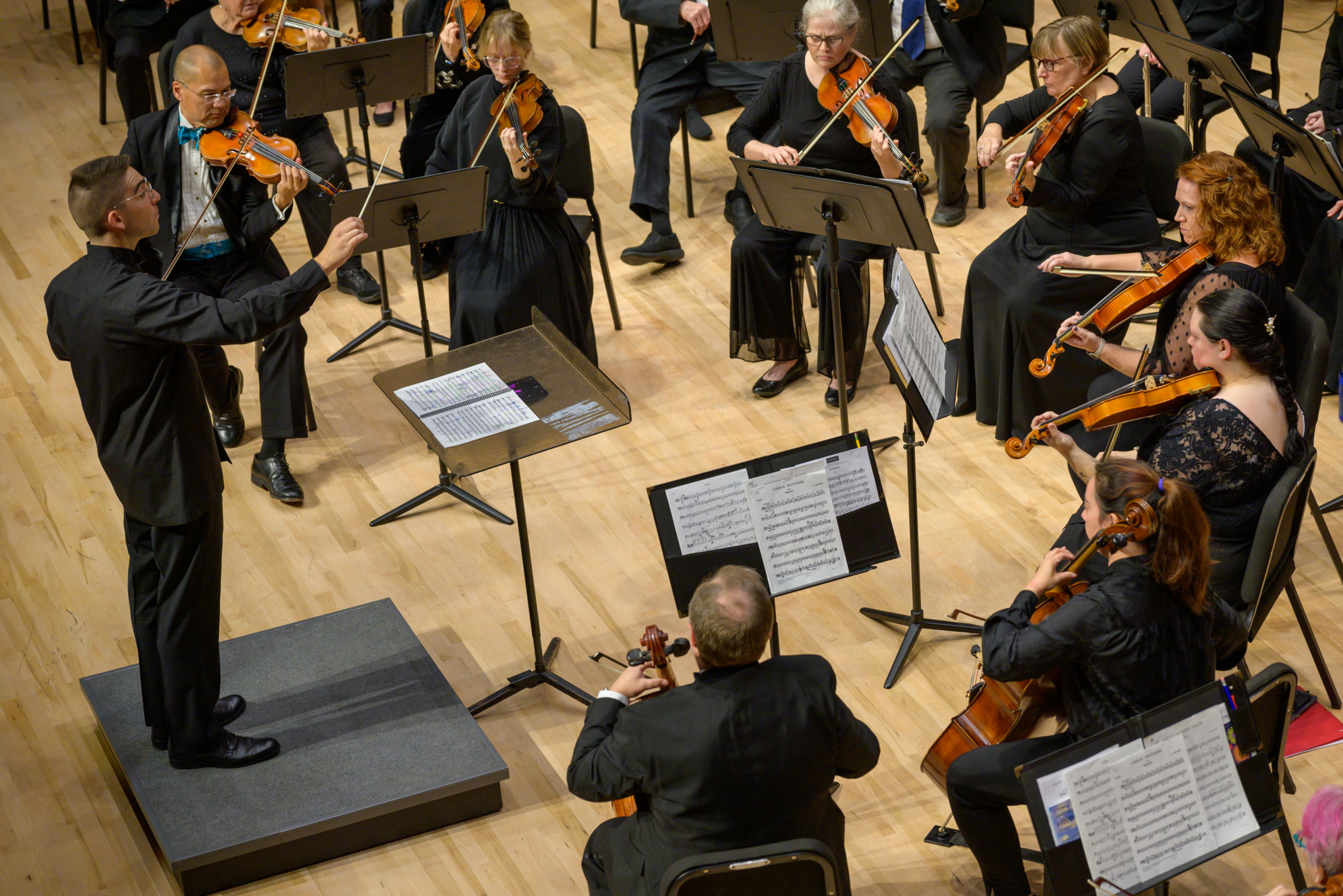 53297582303_56b295a194_o A conductor leads an orchestra of violinists and violists, all dressed in black, performing on a wooden stage with sheet music on stands.